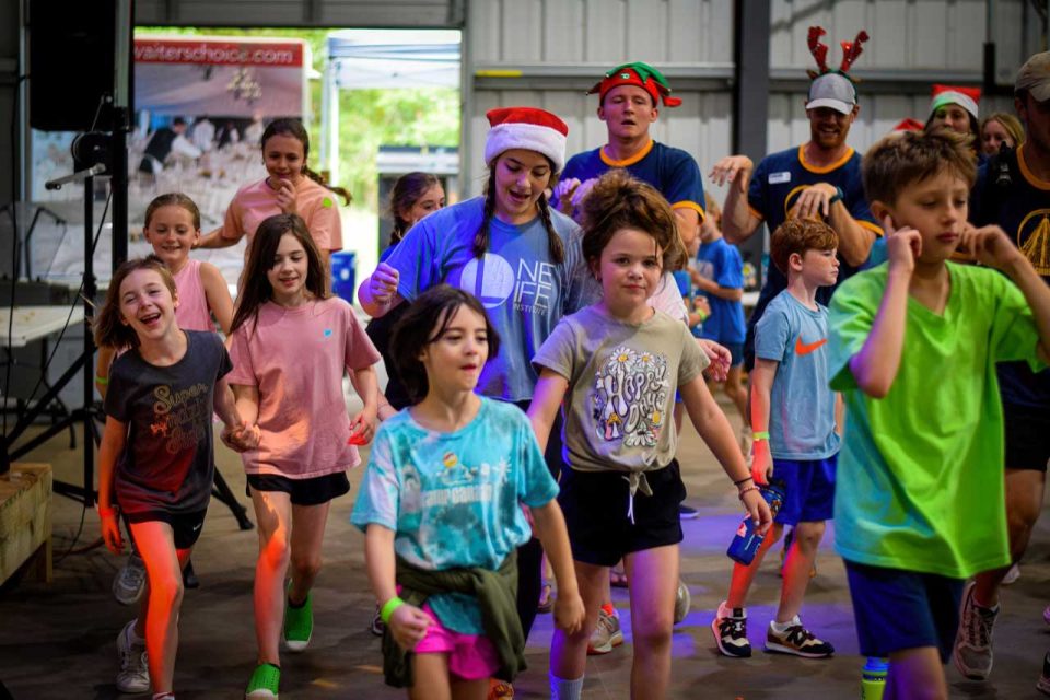 A lively group of children and adults, some wearing holiday-themed hats, participate in an evening activity in the covered basketball court during summer camp.