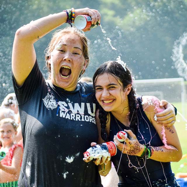 Two girls joyfully spray each other with shaving cream during a sludge war in the sports fields of Camp Canaan. Both are drenched and smiling. The girl on the left holds a shaving cream bottle above her head, while the other girl poses closely, water splashing around them. Other people and campers enjoy the fun summer camp activity.