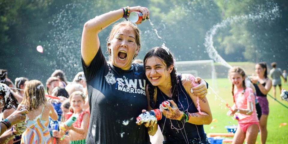 Two girls joyfully spray each other with shaving cream during a sludge war in the sports fields of Camp Canaan. Both are drenched and smiling. The girl on the left holds a shaving cream bottle above her head, while the other girl poses closely, water splashing around them. Other people and campers enjoy the fun summer camp activity.