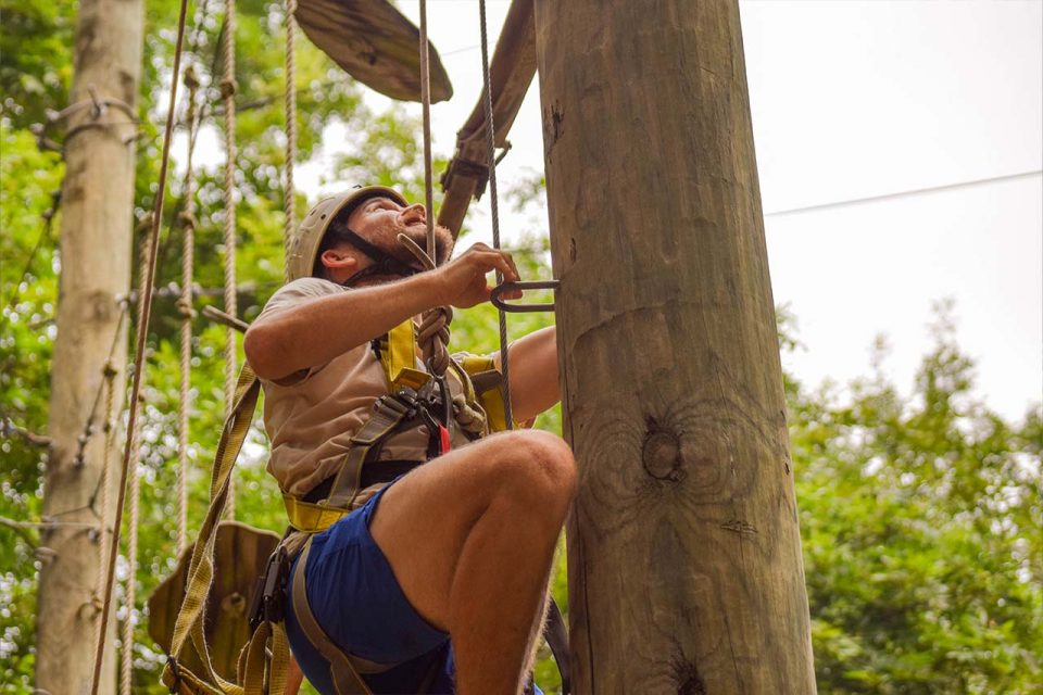 A person wearing a harness and a helmet is seen climbing a tall wooden pole during summer camp at Camp Canaan. The individual appears focused while ascending, and the background shows green foliage.