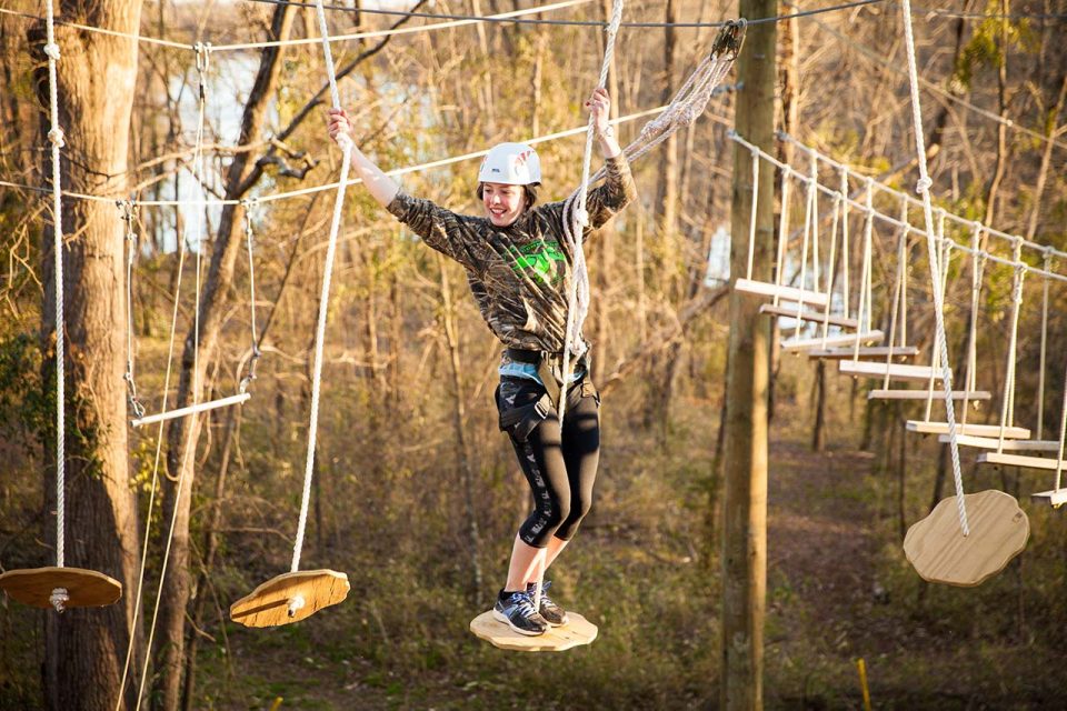 A person with a white helmet and safety harness is navigating a rope obstacle course high above the ground in a wooded area at Camp Canaan. They are standing on a wooden disc while holding onto ropes, smiling and looking ahead, as part of the exhilarating high adventure course.