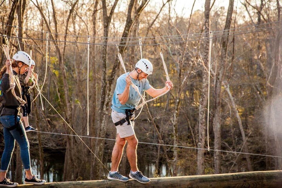 Three people wearing safety helmets and harnesses navigate a high ropes course in a forested area. One person balances on a wooden log while holding onto their harness ropes, with two others following behind on various elements.