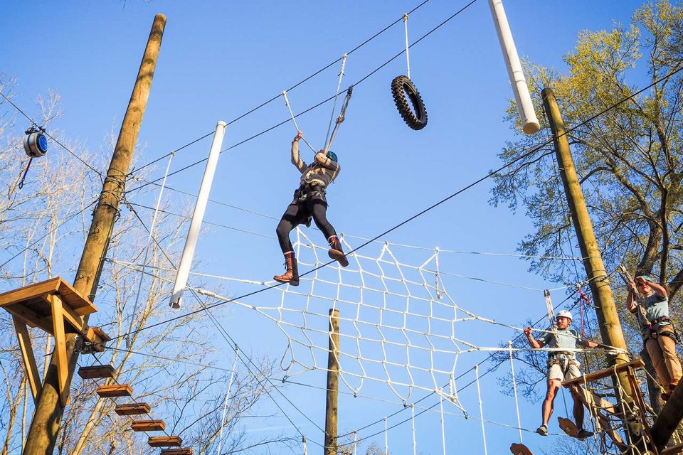 A person wearing a helmet and harness navigates a high ropes course at Camp Canaan, balancing on and gripping various obstacles like ropes, a net, and a tire. Other participants are seen on the course. Trees and a clear sky serve as the background.