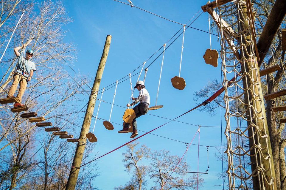 Two people navigate Canaan's high ropes course with various suspended obstacles. Both wear helmets and harnesses for safety. The course, perfect for corporate retreats and summer camp adventures, is set against a backdrop of trees and a clear blue sky.