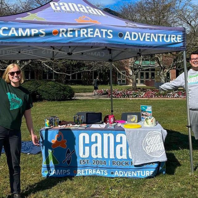 Two people stand under a canopy tent promoting "CAMPS," "RETREATS," and "ADVENTURE." They smile and hold their arms open wide. The table underneath the tent displays pamphlets for summer camp, and branded items. The sky is clear, with trees in the background.