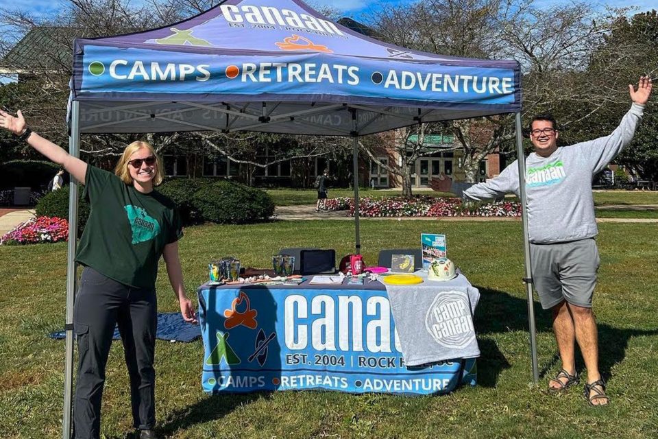 Two people stand under a canopy tent promoting "CAMPS," "RETREATS," and "ADVENTURE." They smile and hold their arms open wide. The table underneath the tent displays pamphlets for summer camp, and branded items. The sky is clear, with trees in the background.