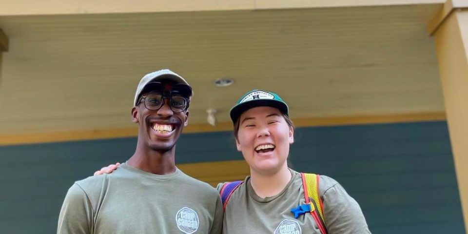 Two people wearing matching green Camp Canaan T-shirts and hats stand outside the Welcome Center.