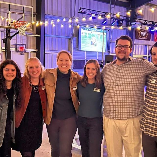 A group of six smiling adults stands closely together in the covered basketball court at Camp Canaan, decorated with string lights, a basketball hoop, and a screen displaying an image in the background. They appear to be at an event, with tables and chairs set up around them.