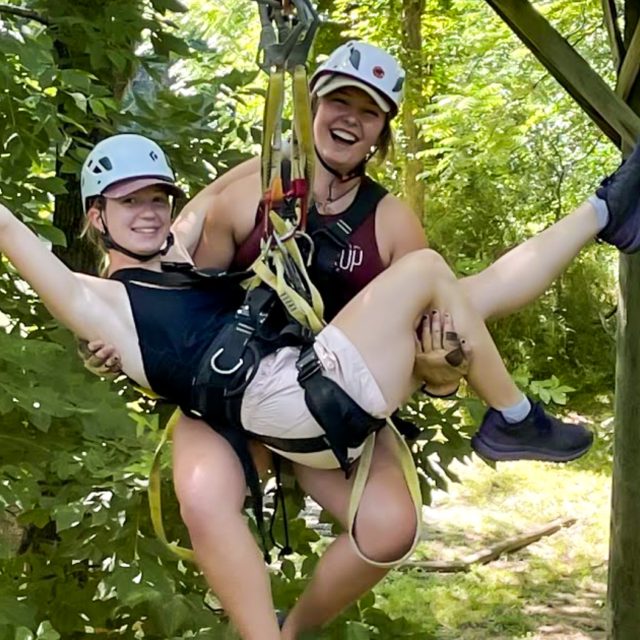 Two people wearing helmets and harnesses smile while ziplining through the lush, green trees at Camp Canaan's summer camp and retreat center. One person is holding onto the other, and they both appear joyful and excited.