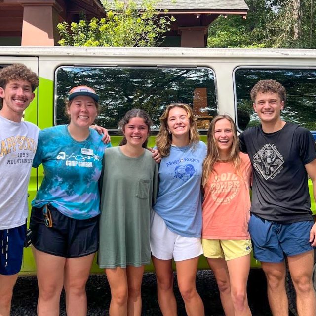 A group of six smiling young adults stand in front of a green van with "Camp Canaan" written on it. Dressed casually in t-shirts and shorts, they look ready for summer camp. Behind them, trees and the multi-purpose building show some of the Camp amenities. It is a sunny day.