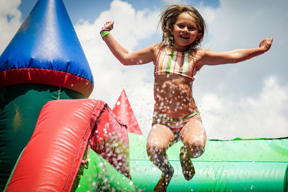 At Camp Canaan's summer camp, a young girl in a striped swimsuit leaps joyfully from a colorful inflatable structure, with water splashing around her. She appears to be playing outdoors on a sunny day, and the sky is visible in the background.