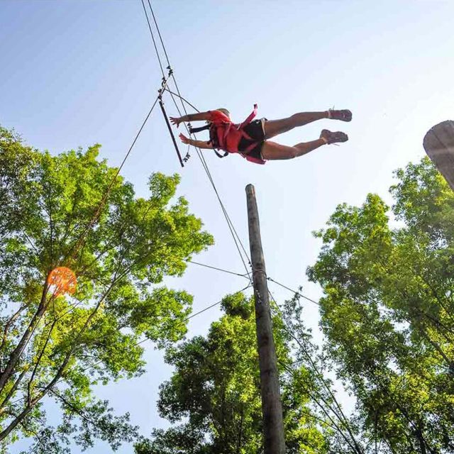 Camper in red shirt and black shorts makes a huge jump on the leap of faith element during summer camp. It appears that they will reach the hanging bar after jumping off of the wooden pole. It is a sunny day.