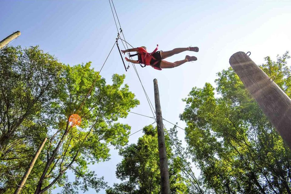 Camper in red shirt and black shorts makes a huge jump on the leap of faith element during summer camp. It appears that they will reach the hanging bar after jumping off of the wooden pole. It is a sunny day.