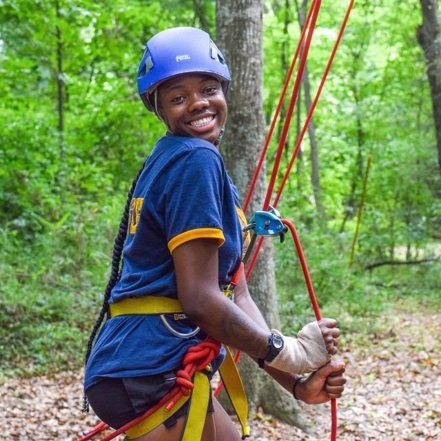 An adventure guide wearing a blue helmet and blue shirt with yellow accents is smiling while holding red climbing ropes in a wooded setting at Camp Canaan. The background shows trees and greenery.