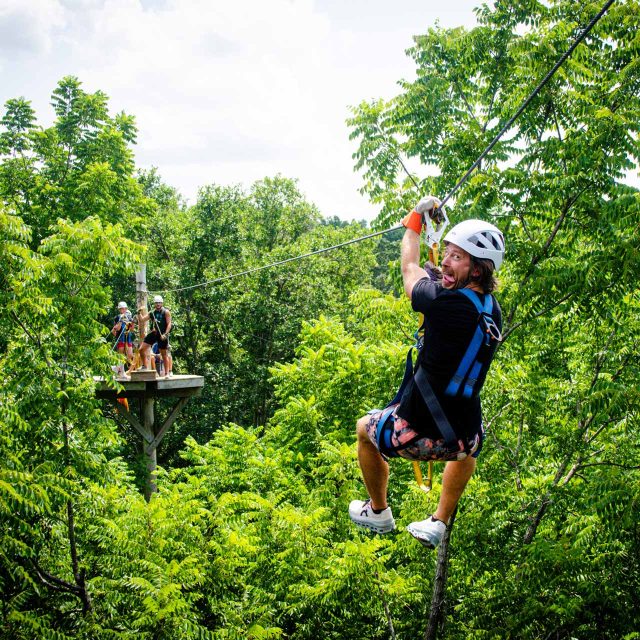 A person in a white helmet and harness is smiling while ziplining through lush green trees at a summer camp in Rock Hill, SC. In the background, a group of people stand on a wooden zipline canopy tour platform among the trees.