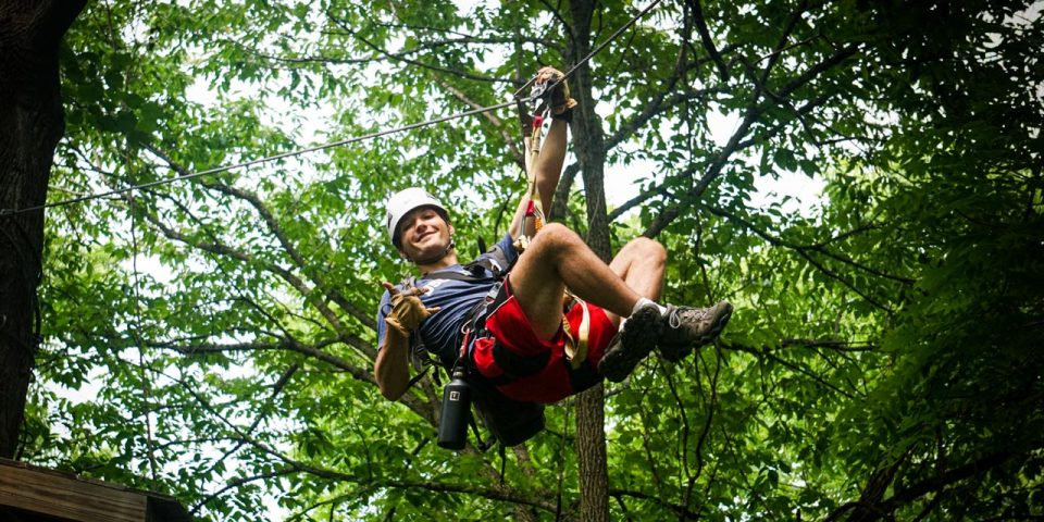 A person wearing a helmet and harness smiles and gives a thumbs-up while ziplining through a lush, green wooded area at Camp Canaan.